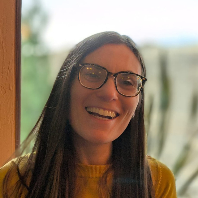 Caucasian female, smiling, with long brown hair, wearing glasses and a yellow sweater.