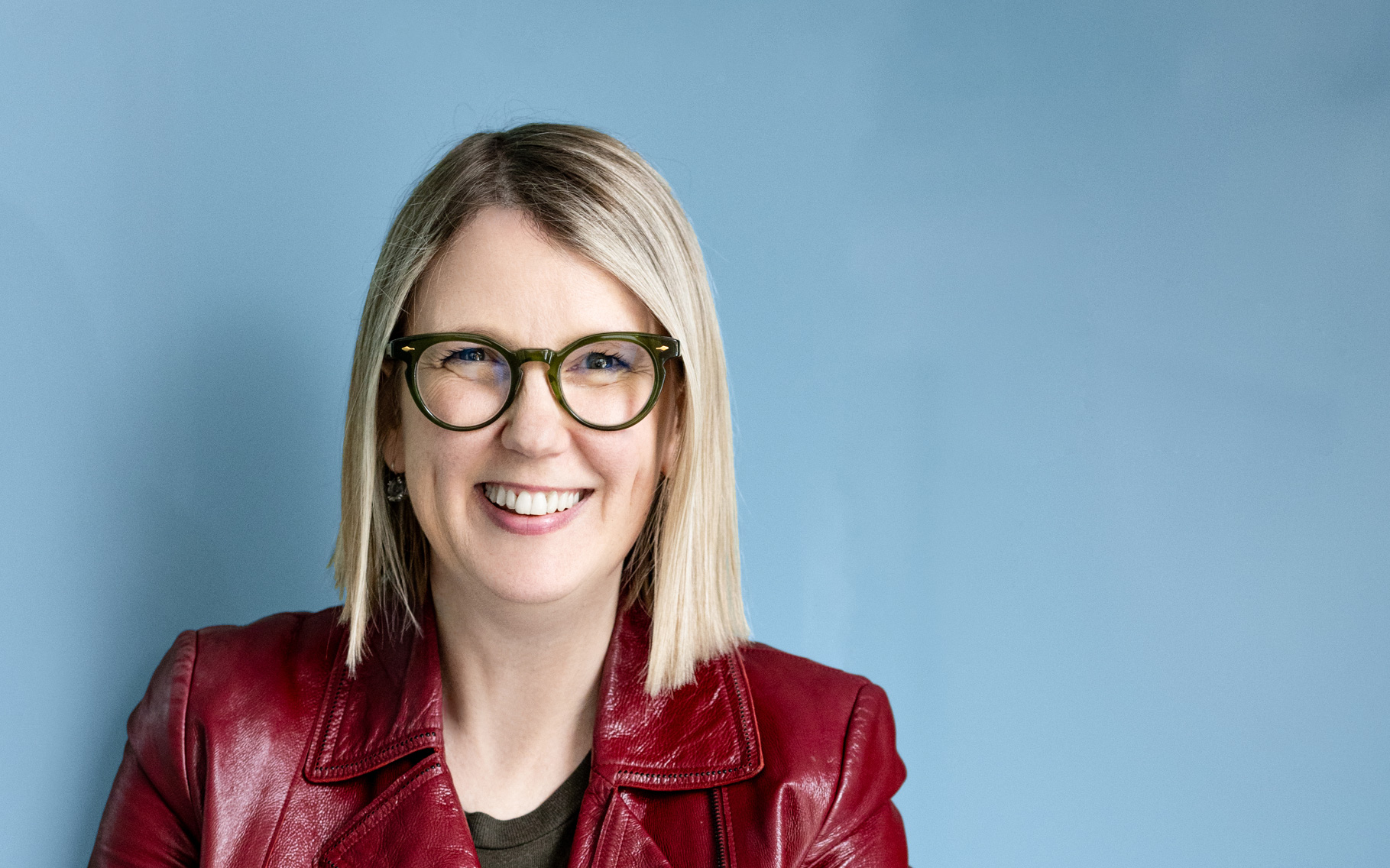 Jenny, a woman with straight blonde hair and green-framed glasses, smiling warmly while wearing a red leather jacket over a dark top, posed against a light blue background.