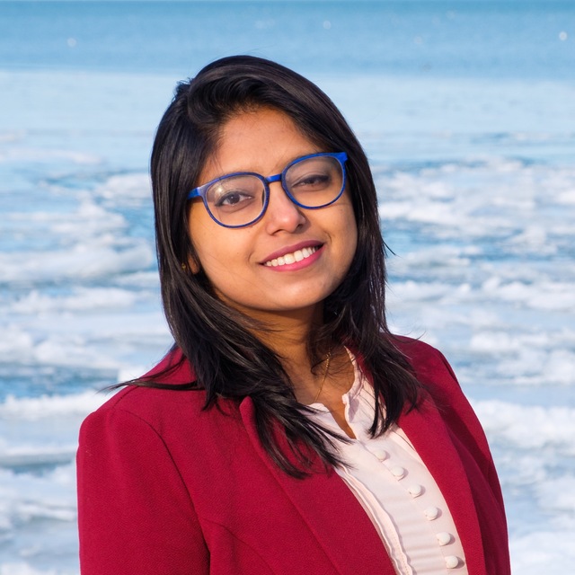 Maitraye, a Brown woman with black shoulder-length straight hair, is smiling at the camera. She is wearing round, blue-rimmed glasses and a red blazer over a pink colored shirt.