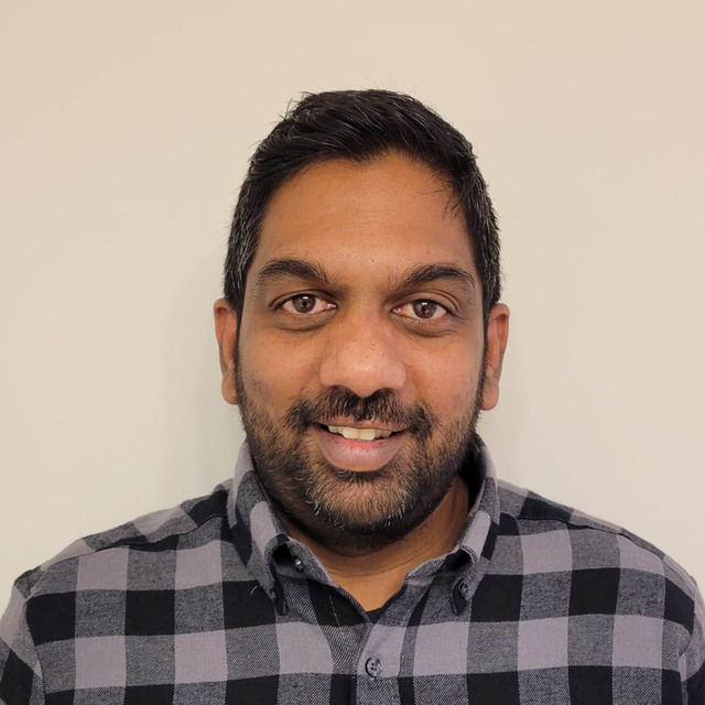 A selfie of Roshan, a brown man with short black hair and a beard. He is wearing a black and gray checkered shirt. 