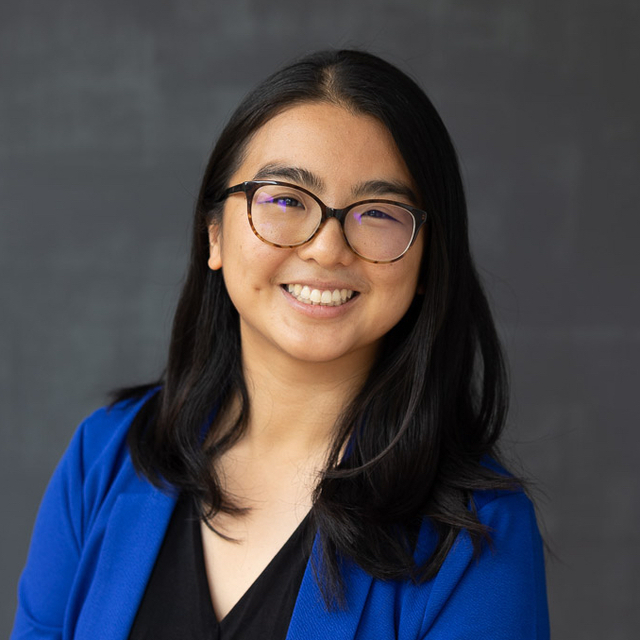 Headshot of Emily, an Asian American woman. In this photo, she has medium length black hair and is wearing brown glasses, a blue blazer, and a black V-neck top.
