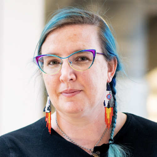 A picture of Kat, a white woman with blue-tinted hair in a braid, wearing colorful beaded earrings and glasses.