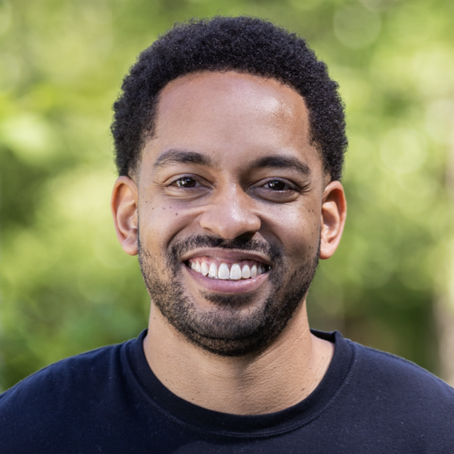 A picture of Martez, a Black man with a short, black afro and a stubble beard wearing a black t-shirt and smiling while standing outside in front of trees blurred in the background.