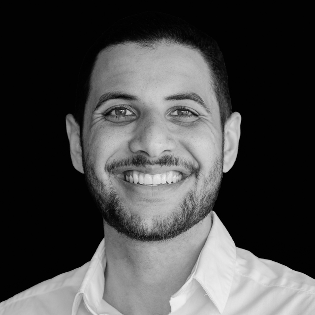 Black and white studio portrait of Oliver Alonzo with a big smile, short black hair and beard, wearing a white shirt.