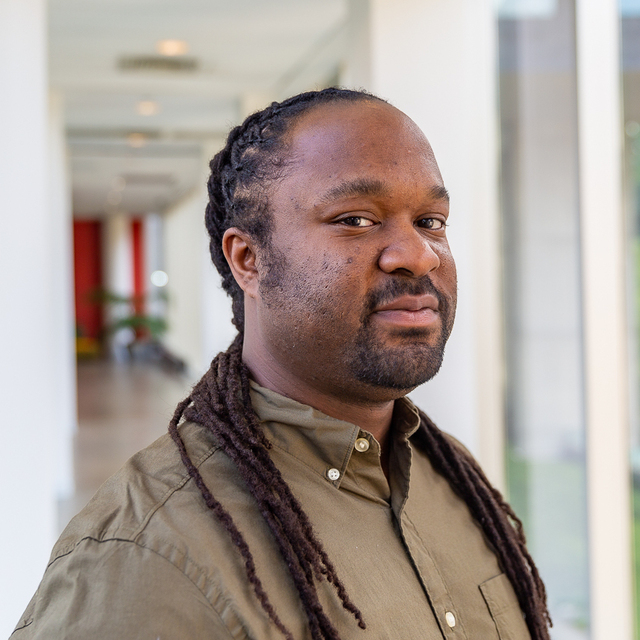 A man with long dreadlocks wearing a green button-up shirt. The individual has a calm, confident expression and is positioned in what appears to be a modern, well-lit indoor space with glass walls and a hallway.