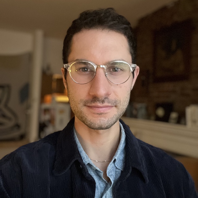 Headshot of Willie, a white man with short black hair and stubble. He is smiling, wearing silver glasses, a sliver necklace, and layered blue shirts.